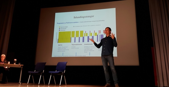 En man står på en scen och föreläser, en projektorskärm i bakgrunden